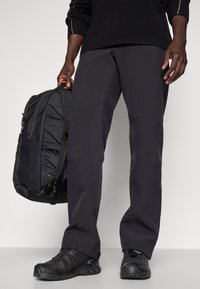 Calças de caminhada pretas com corte reto, combinadas com uma mochila preta com painéis texturizados e correias ajustáveis, usadas com calçado preto.