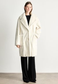 Palton din blană sintetică în crem, cu guler larg, design dublu-îmbăcat și buzunare mari, asortat cu pantaloni negri evazați.