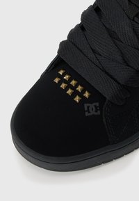 Zapatilla de ante negra con un cordón grueso negro, tachuelas de metal dorado en la puntera y un logotipo texturizado negro en el lateral.