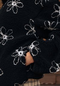 Sort fleece-pullover med hvid blomsterbroderi. Har en struktureret overflade og løstsiddende ærmer med blomsteromrids ved manchetterne.