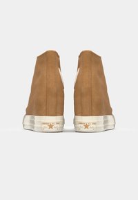 Brune suède høyhælte sneakers med en hvit logo på hælen og en teksturert gummisåle. Merkevarekjennskap på siden og baksiden.