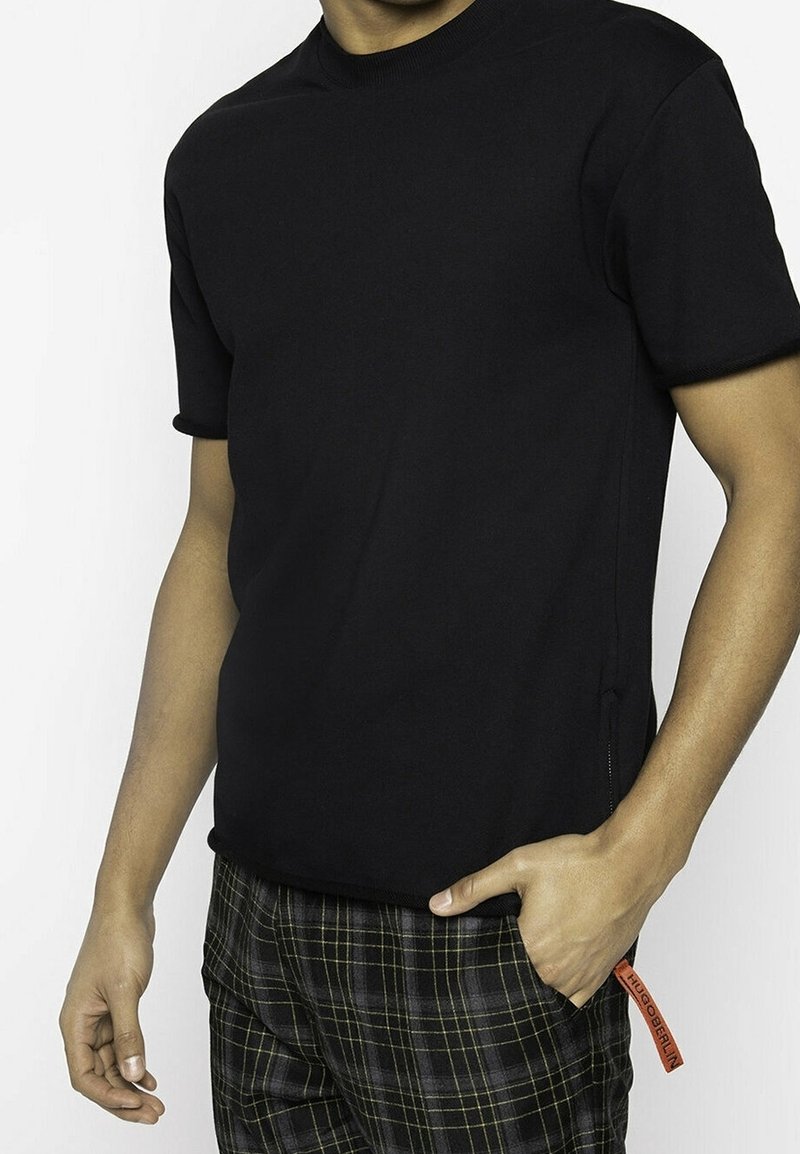 Homme portant un t-shirt noir uni à manches courtes et un pantalon à carreaux noir avec une étiquette rouge indiquant "HUGO BERLIN" dans la poche.