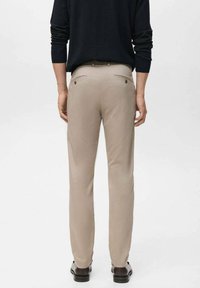 Beige Hose mit slim Fit, aus weichem Stoff gefertigt. Verfügt über zwei Gesäßtaschen mit Knöpfen und sauberen Nähten. Kombiniert mit einem dunklen Oberteil.