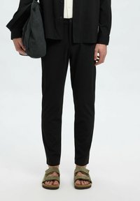 Schwarze, leichte Hose mit Slim Fit, kombiniert mit olivgrünen Sandalen mit zwei Riemen. Eine graue Schultertasche wird in einer Hand gehalten.
