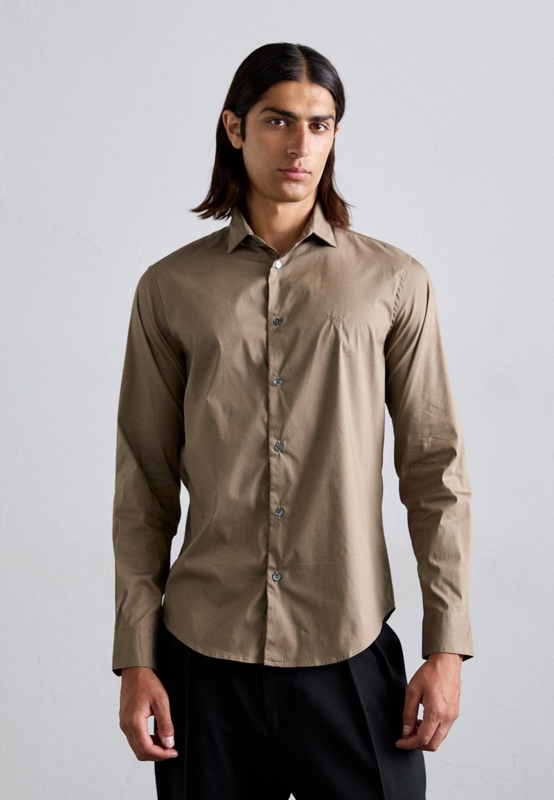 Chemise à manches longues marron en tissu lisse, avec un col classique, une fermeture à boutons à l'avant et un petit logo brodé sur la poitrine.