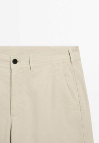 Pantalon beige avec passants de ceinture, poches avant, bouton noir visible et détails de coutures sur fond blanc uni.