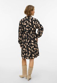 Czarna sukienka z beżowym wzorem liści, długie rękawy i marszczona talia. Modelka nosi jasnobeżowe kozaki do kolan. Gładka faktura, casualowy styl.