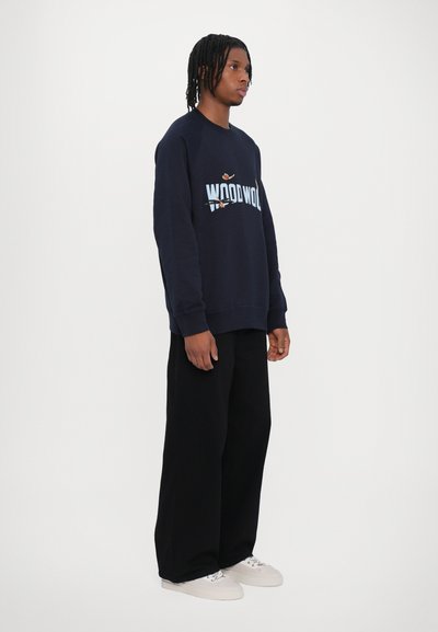 Hanorac bleumarin cu guler rotund, cu logo-ul „WOOD WOOD” în bleu deschis, asortat cu pantaloni negri foarte largi și adidași albi.