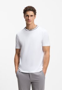 Άνδρας που φοράει λευκό κοντομάνικο μπλουζάκι με τη λέξη «BOSS» στον γιακά και ανοιχτό γκρι παντελόνι, στέκεται με τα χέρια στις τσέπες μπροστά σε λευκό φόντο.