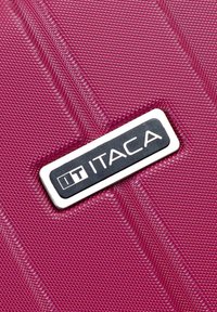 Maleta texturizada de color púrpura con una etiqueta de logo. La superficie tiene un patrón en relieve y un diseño angulado. Presenta un acabado plateado brillante alrededor del logo.