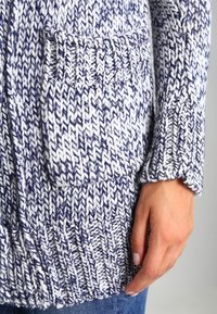 Marinblå och vit stickad kardi med ett texturerat mönster, utrustad med en framficka och ribbade ärmslut. Mjuk garn och lös passform.