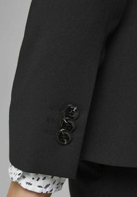 Ärmen på en svart kavaj med tre svarta knappar, texturerat tyg och en vit skjortmanschett med ett litet lövmönster.