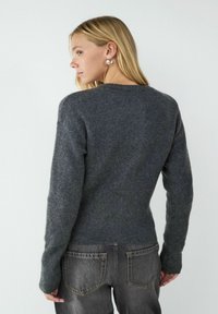 Dunkelgrauer Pullover mit langen Ärmeln, geripptem Saum und strukturiertem Stoff. Kombiniert mit dunklen Jeans, die eine schmale Passform und minimale Details zeigen.
