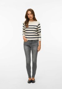 Femme aux longs cheveux bruns portant un pull à rayures crème et marine avec un écusson smiley, un jean skinny gris et des ballerines noires.