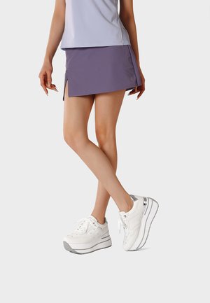 Donna che indossa una gonna asimmetrica lilla con spacchi laterali, sneakers bianche con platform e un top senza maniche di colore chiaro, in piedi con le gambe incrociate.