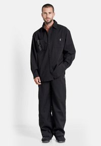 Man met kort haar en een baard, gekleed in een losse zwarte gestructureerde shirt en broek, met zwarte schoenen, en een zonnebril in de zak van zijn shirt gestoken.