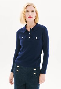 Marineblauwe gebreide poloshirt met kraag, voorzien van twee borstzakken en zilveren knopen. Lange mouwen en een getailleerd ontwerp.