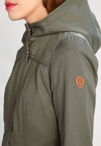 Oliwkowo-zielona kurtka z kapturem o teksturowanym wykończeniu, z zapięciem na zatrzaski, marszczonymi rękawami oraz brązowym skórzanym akcentem na ramieniu.