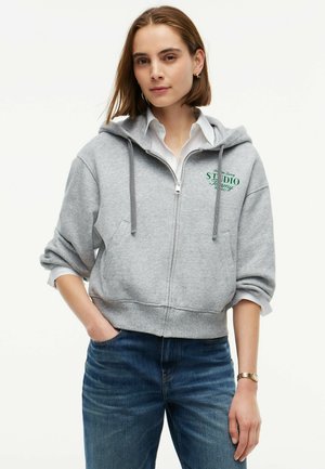 Kvinde iført grå hættetrøje med lynlås over hvid skjorte og blå jeans med den ene hånd i lommen mod en ensfarvet baggrund.