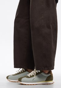 Zapatillas de deporte azul claro con acentos de gamuza verde, bordes festoneados y cordones blancos, combinadas con pantalones sueltos de color marrón oscuro.