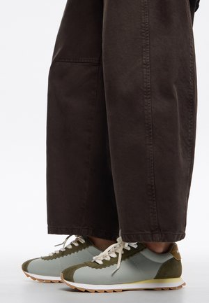 Zapatillas de deporte azul claro con acentos de gamuza verde, bordes festoneados y cordones blancos, combinadas con pantalones sueltos de color marrón oscuro.