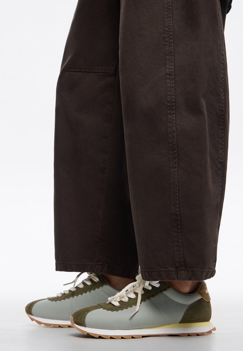 Zapatillas de deporte azul claro con acentos de gamuza verde, bordes festoneados y cordones blancos, combinadas con pantalones sueltos de color marrón oscuro.