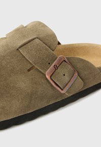 Sandál z olivovo zelenej semišovej kože s hnedou kovovou prackou, plochou čiernou podrážkou a prírodnou stielkou. Má jeden remienok cez priehlavok.