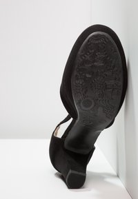 Chaussure à talon en suède noir positionnée semelle vers le haut, montrant une semelle extérieure en caoutchouc à motif floral et un talon bloc sur fond blanc.