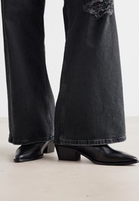 Botas de tornozelo pretas com salto alto, ponta afilada e acabamento em couro texturizado, combinadas com jeans pretos de perna larga desgastados, com bordas desfiadas.