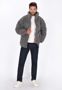Mand, der står selvsikkert iført en grå fleecejakke, hvid skjorte, mørke jeans og hvide sneakers foran en ensfarvet hvid baggrund.