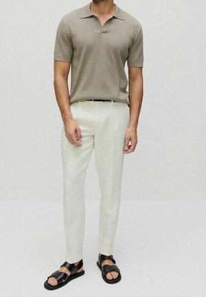 Homme portant un polo beige à manches courtes, un pantalon blanc sur mesure et des sandales noires à bouts ouverts, debout devant un fond clair uni.
