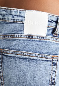Ljusblå denimjeans med synlig sömnad, utrustade med en vit läderlapp märkt "BOSS" bak vid midjan. Klassisk femfickdesign.