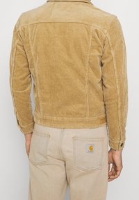 Giacca di velluto a coste di colore marrone chiaro con una superficie testurizzata, polsini con bottoni e un dettaglio sul retro. Abbinata a jeans beige chiaro con etichetta.