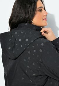 Žena s tmavými vlasy upravuje kapuci černé bundy s jemným vzorem tmavě šedých hvězd, stojící bokem na bílém pozadí.