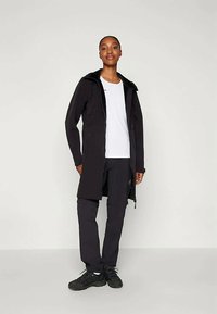 Black long coat with a hood, worn over a white shirt, paired with dark trousers and black sneakers, standing against a light background.