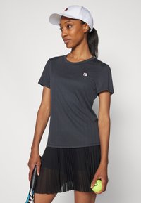 Σκούρο γκρι αθλητικό μπλουζάκι με κοντό μανίκι και κατακόρυφες ρίγες, συνδυασμένο με πλισέ μαύρη φούστα. Λευκό καπέλο με λογότυπο και ρακέτα τένις.