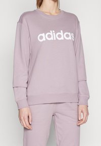 Svetle fialová mikina vyrobená z měkké látky, na hrudi s bílým logem "adidas". Pružné manžety a lem pro přiléhavý vzhled.