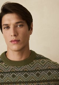 Fiatal férfi, rövid sötét hajjal és kék szemekkel, zöld mintás kötött pulóvert visel, sima bézs háttér előtt.