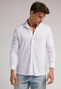 Camisa blanca de manga larga hecha de una tela suave con botón frontal, cuello clásico y un ajuste slim. Estilizada con jeans de mezclilla azules.