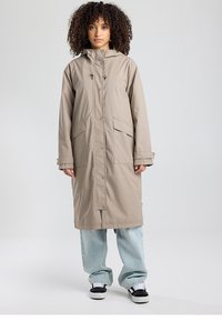 Manteau imperméable beige avec capuche, boutons-pression et grandes poches plaquées. Coupe ample avec manches resserrées aux poignets, porté sur un jean bleu clair et des baskets.