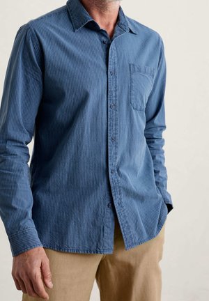 Blau gestreiftes Langarmhemd mit Button-Down-Kragen und einer Brusttasche, aus einem strukturierten Stoff. Getragen mit beigen Hosen.