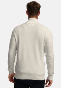 Man die van achteren is gezien, draagt een witte gebreide trui met structuur, geribbelde kraag, manchetten en zoom, gecombineerd met een donkere broek tegen een witte achtergrond.