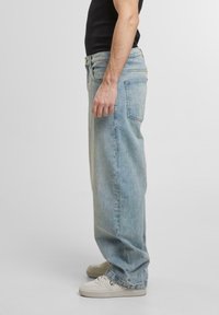 Jeans in denim azzurro chiaro con una vestibilità rilassata, caratterizzati da un alto girovita, tasche laterali e dettagli sbiaditi discreti. Indossati con sneakers bianche.