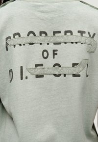 Πίσω μέρος ανοιχτόγριζου ενδύματος με μαύρο κείμενο "PROPERTY OF D.I.ESEL" που καλύπτεται εν μέρει από υφασμάτινες λωρίδες με υφή και ραφές.