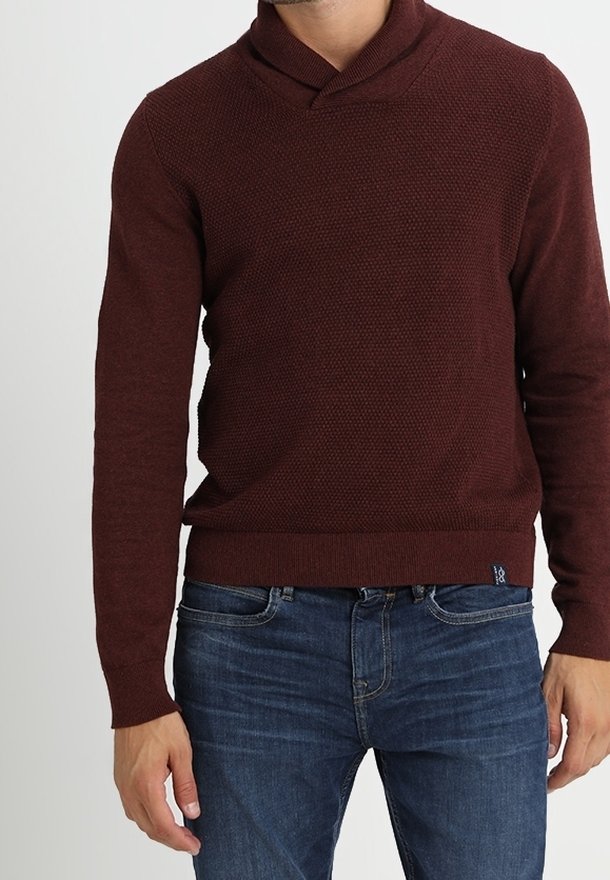 Homme portant un pull à col châle texturé rouge foncé associé à un jean bleu foncé, debout devant un fond uni.
