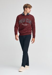 Bordeaux sweatshirt med texten "POLO CLUB", bärs över en blå skjorta med krage, parat med svarta byxor och ljusa sneakers. Modellen går.