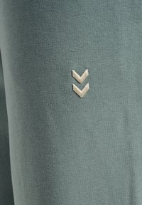 Grüne Stoffleggings mit einer glatten Textur und einem kleinen, cremefarbenen Chevron-Logo, das nahe der unteren Seite gestickt ist.