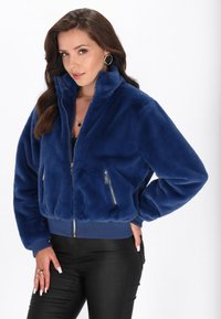 Giacca in pelliccia sintetica di colore blu navy con collo alto, chiusura anteriore con zip e polsini a costine. Presenta due tasche frontali con zip e una vestibilità corta.