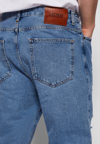 Světlé modré džínové kalhoty s texturovaným provedením, designem pěti kapes a hnědým koženým štítkem s nápisem "2YDNM" na zadní části pasu.