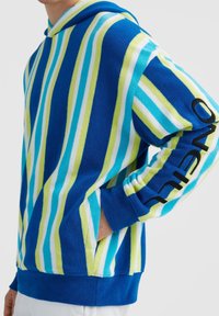 Personne portant un sweat à capuche rayé bleu, vert, blanc et turquoise avec une poche avant et un texte noir sur la manche.